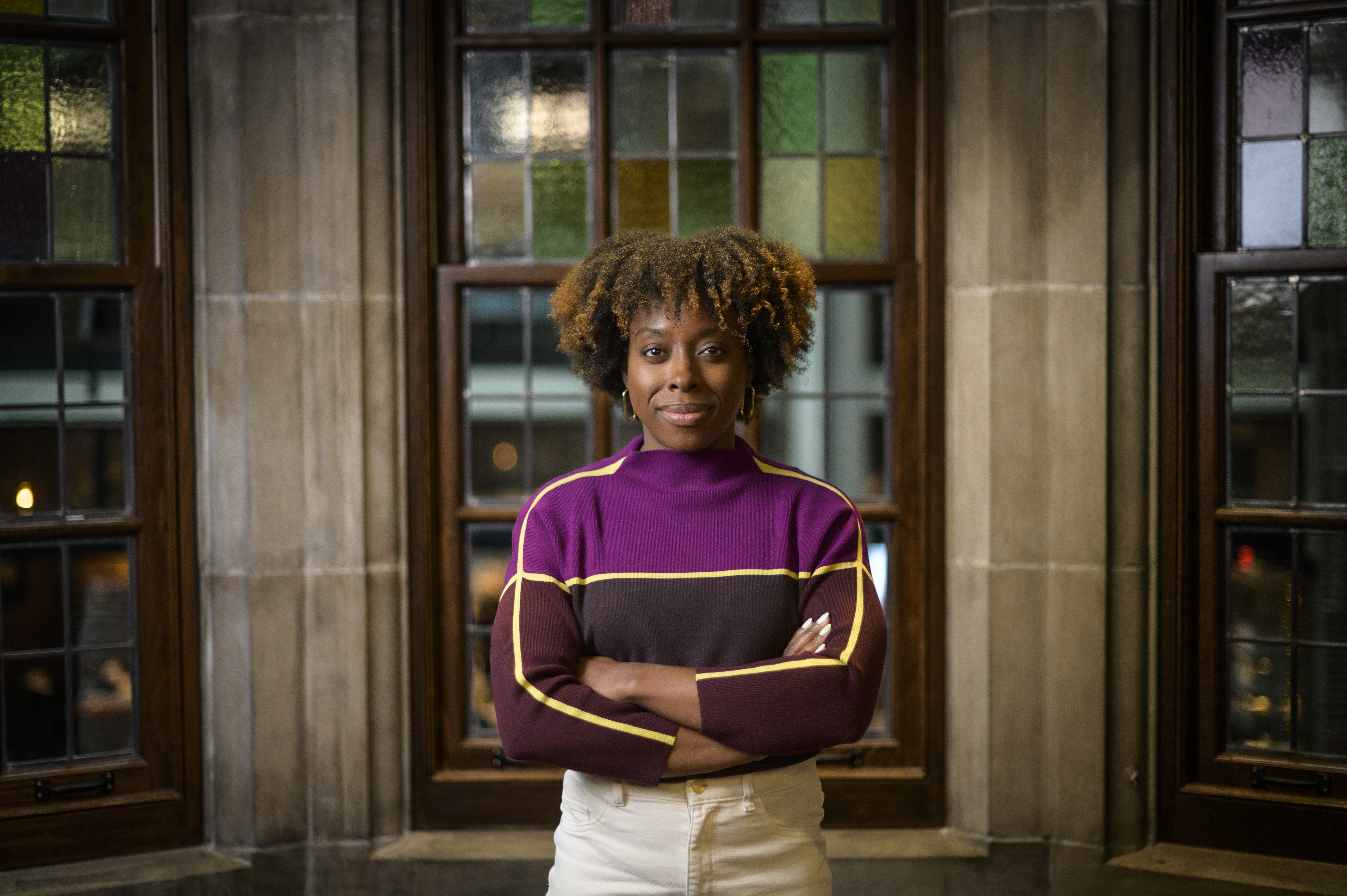 Robin poses in the Michigan Union atrium, with old stone walls and simple stained glass windows framed in wood, while wearing a vibrant shirt of hues of purple with yellow lines.