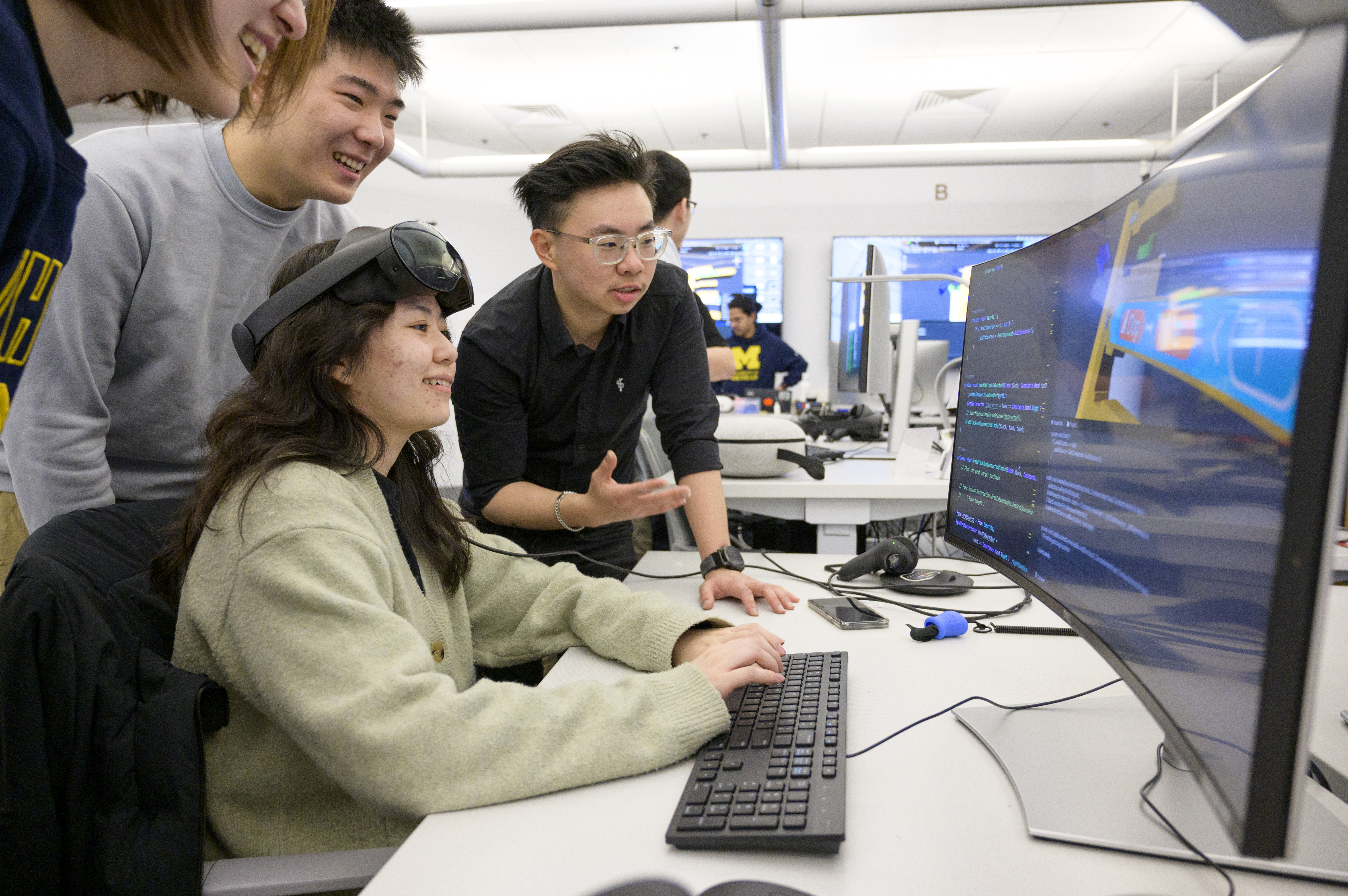 Students gathered around a large computer monitor and talking. The student sitting at the keyboard is wearing VR goggles on top of their head.