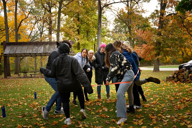 A group of people outside, standing in a circle with linked arms, attempts to get out of a rope around their legs. There are fall trees in the background and leaves on the ground.