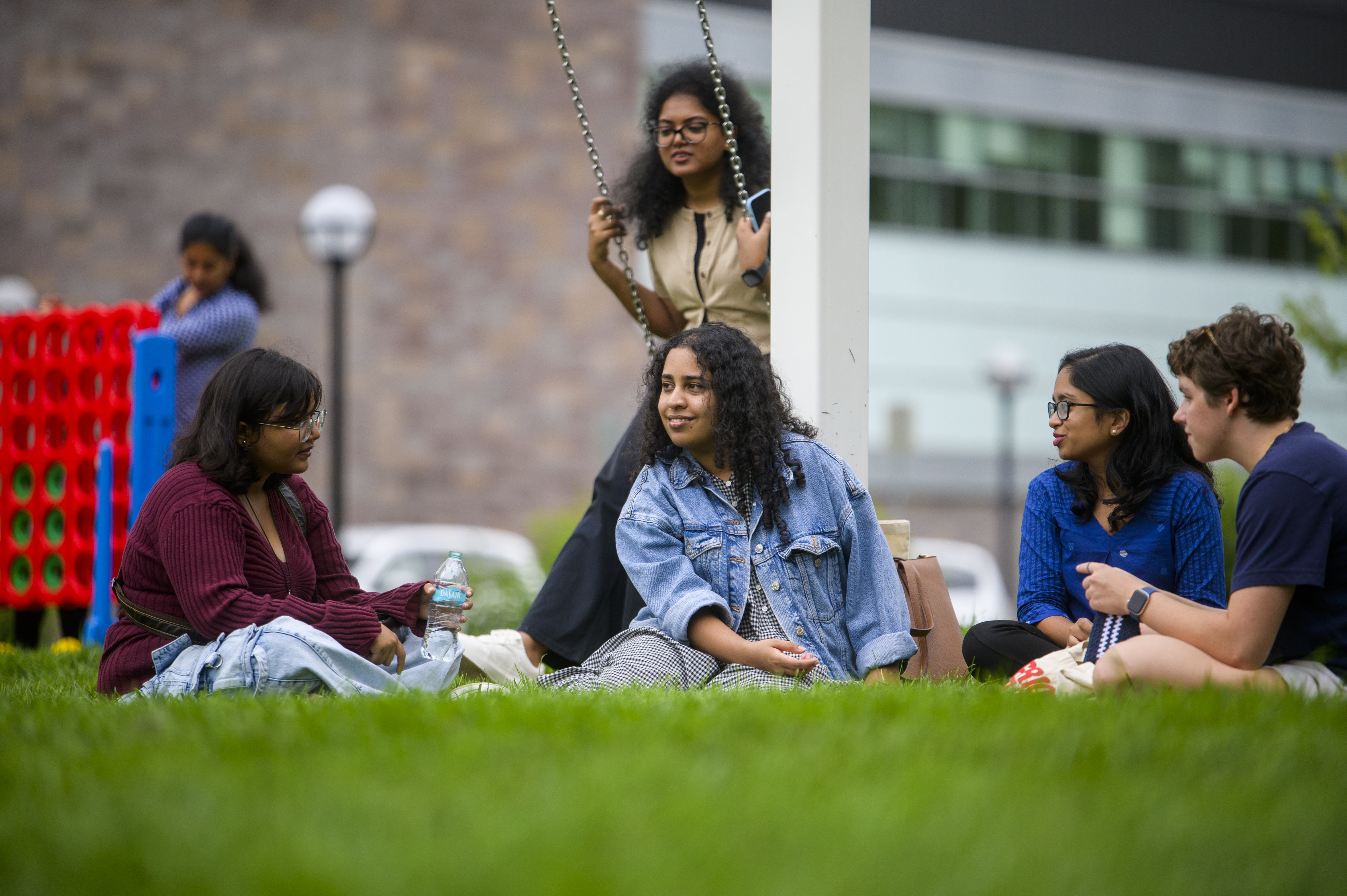 Four students sitting in the grass and chatting. The person behind them is sitting on a swing.