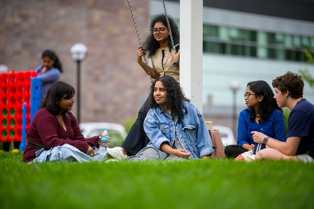 Four people sit on the grass and chat. A person behind them is sitting on a swing.
