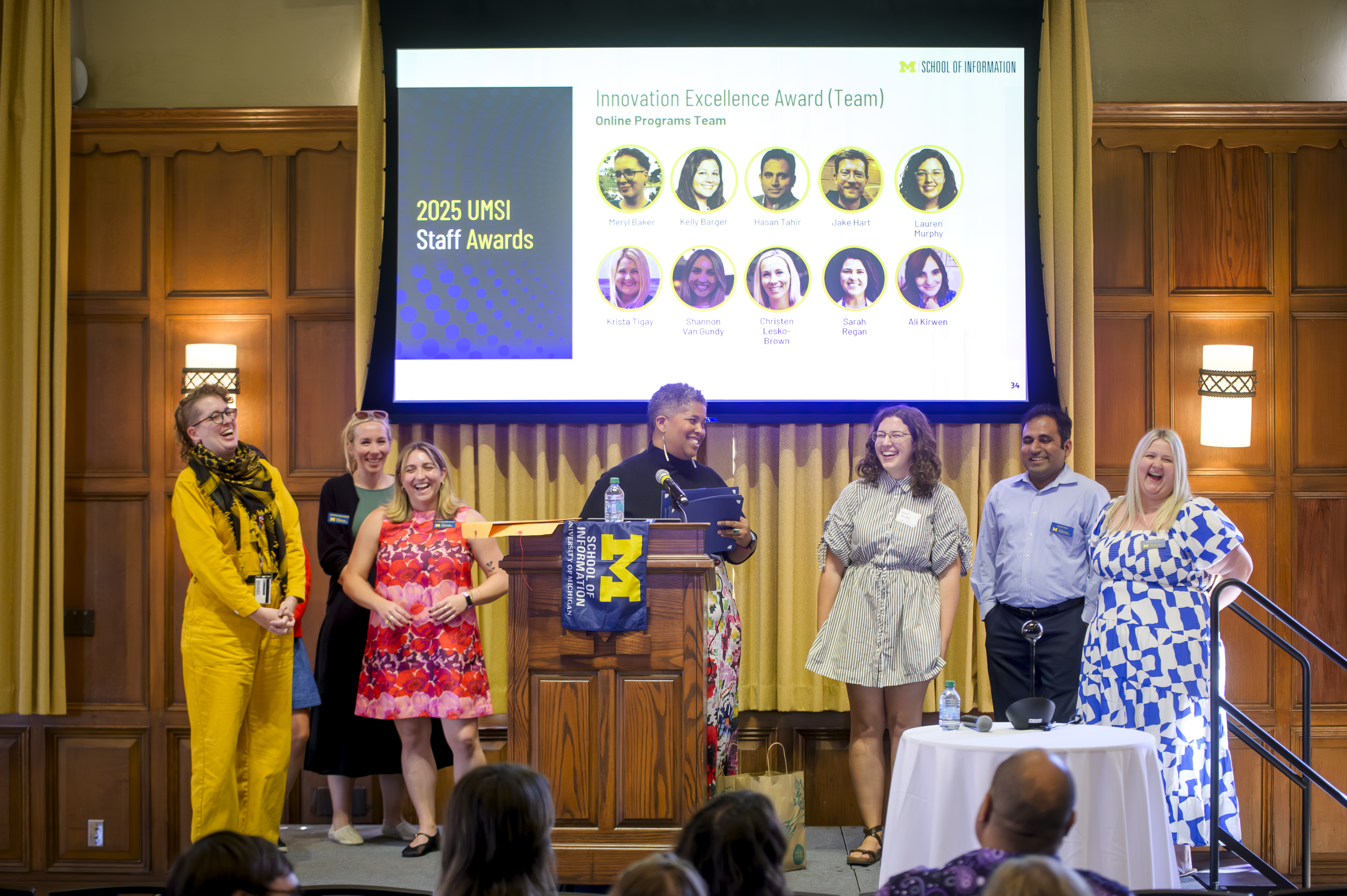 A group of 7 smiling people stand together on a stage behind a podium. The projector screen behind them says, "2025 UMSI Staff Awards. Innovation Excellence Award (Team): Online Programs Team." There are 10 headshots below the text.