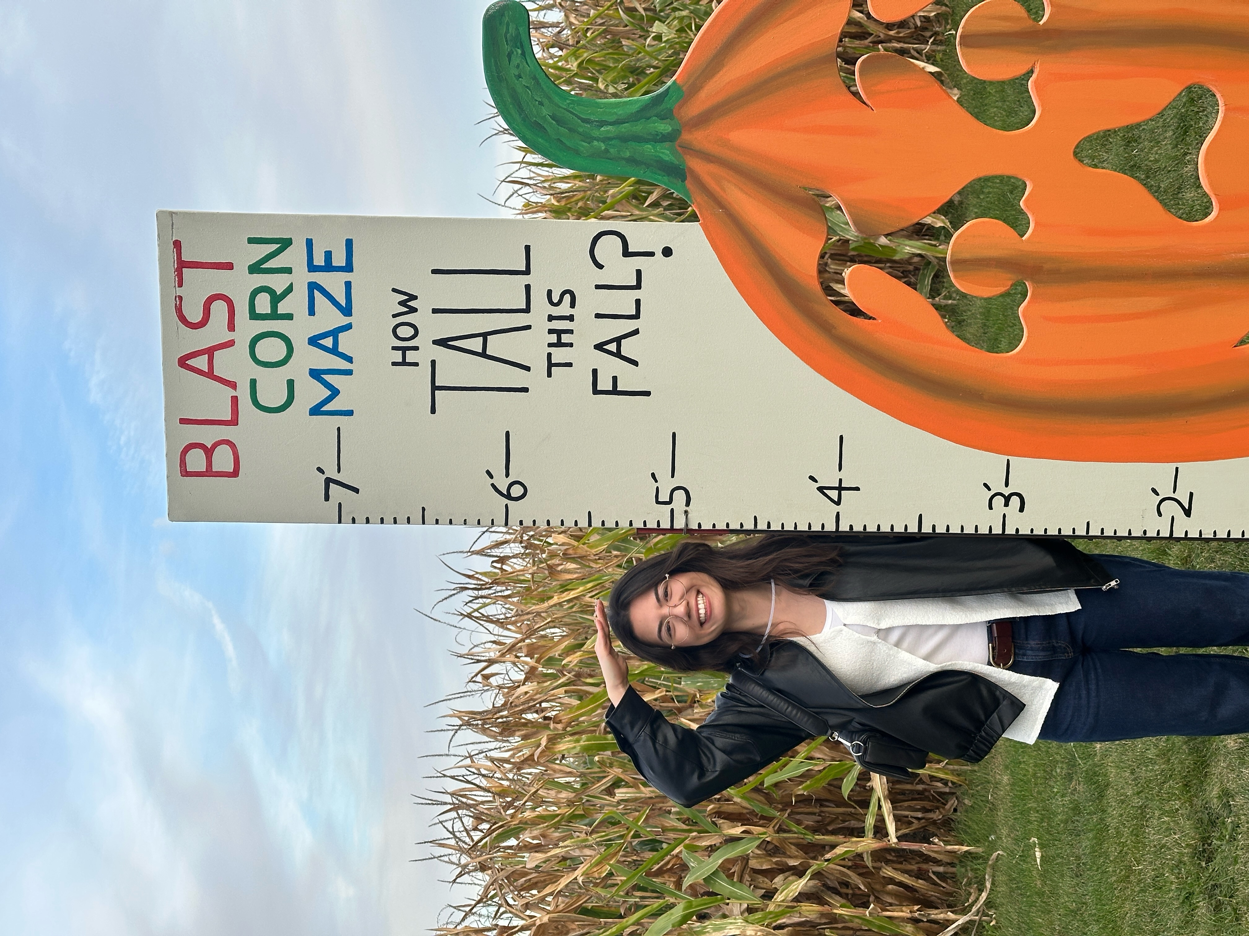 Chandra Behr standing in front of a corn field next to a board that measures 7 feet and asks "How tall this Fall?" Chandra is about 5'5". The signs says, "Blast Corn Maze."