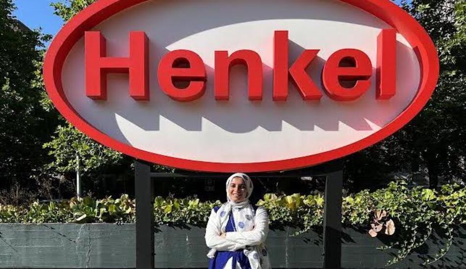 University of Michigan Master of Applied Data Science degree program graduate Rania El-Shenety smiles and stands with her arms crossed in front of her in front of a large sign for the company Henkel, where she was hired as a data scientist. 