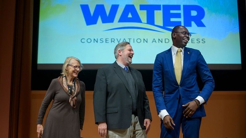 Elizabeth Yakel, Cliff Lampe and Lt. Gov. Garlin Gilchrist II smile on stage in front of a screen projecting "UMSI theme year 2023-24: Water conservation and access"