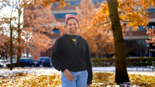 An image of Christine Carethers standing on campus surrounded by fall foliage, wearing a U-M t-shirt and a red bandana
