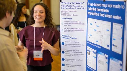 A student speaks with an Expo guest in front of a trifold poster about expanding clean water access for the homeless community.
