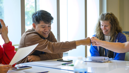 A student smiles while fist bumping another student who is just out of view. They are seated at a table with worksheets in front of them and sunny windows behind them.