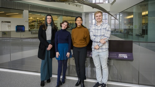 Madison Haack, Julia Cave Arbanas, Cameron Robertson and Austin Book pose on the second level of a museum with a life-size model of a prehistoric pterosaur visible behind them