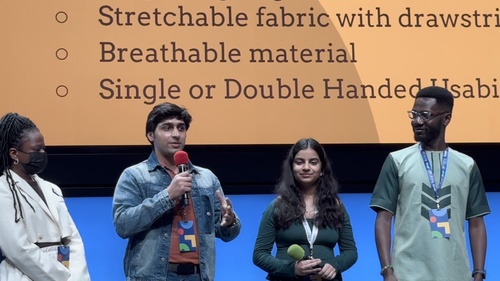 Four students stand on a stage in front of a digital slide. One speaks thoughtfully into a microphone 
