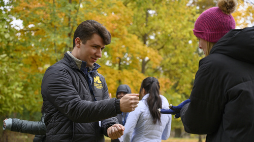 Two people play rock, paper scissors outside. They are wearing warm coats and the trees behind them are changing color.