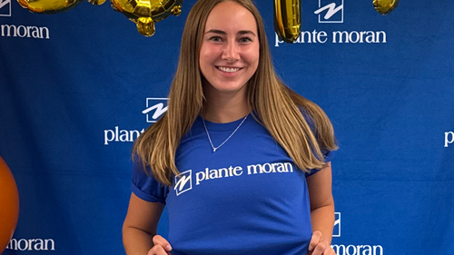 A student wearing a shirt with the Plante Moran logo standing in front of a background with the Plante Moran logo on it.