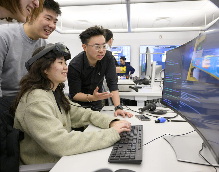 Students gathered around a large computer monitor and talking. The student sitting at the keyboard is wearing VR goggles on top of their head.