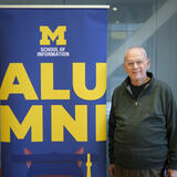 A person stands next to a banner that says Alumni with the UMSI logo.
