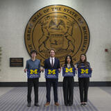 Four students holding School of Information flags stand in front of a wall with a large State of Michigan seal on it.