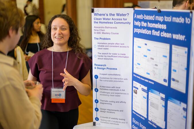 A student speaks with an Expo guest in front of a trifold poster about expanding clean water access for the homeless community.