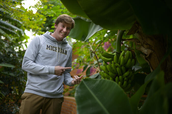 A student wearing a University of Michigan hoodie points at a plant in a greenhouse.