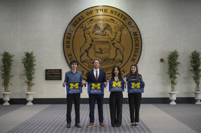 Four students holding School of Information flags stand in front of a wall with a large State of Michigan seal on it.