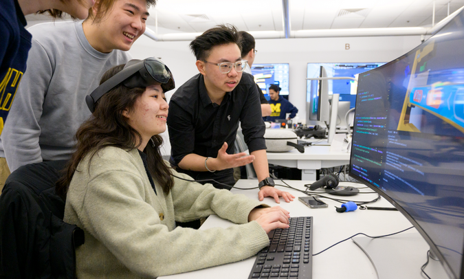 Three students gathered around a computer monitor. The student at the keyboard has VR goggles pushed up on their forehead.