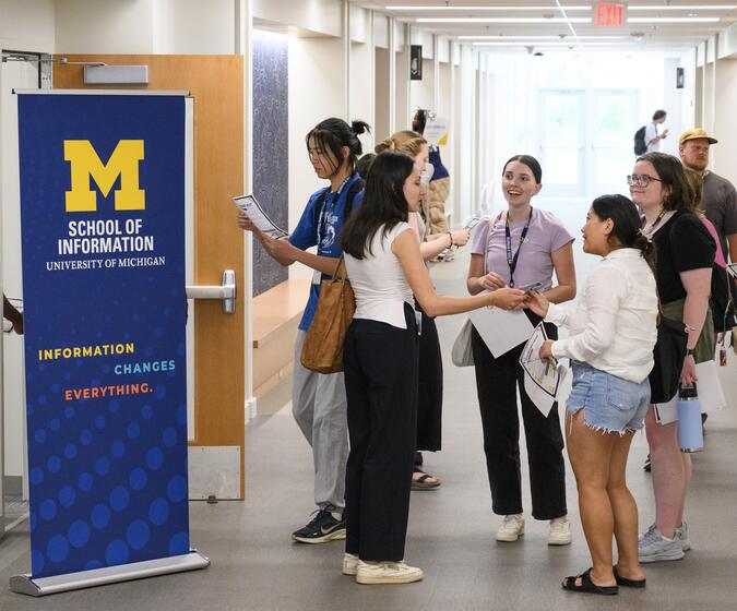 Students congregate, smiling, in a brightly lit hallway with a UMSI pull-up banner 