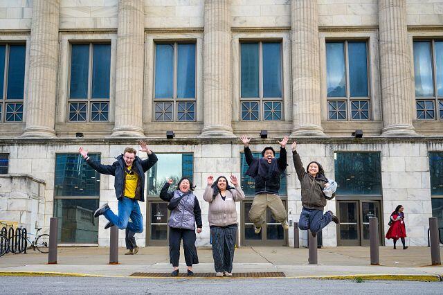 Five people jumping up in the air in front of a building.