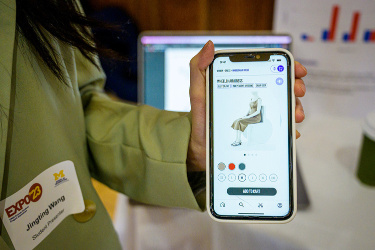 A student holds a smartphone displaying the prototype of the app their team designed. The screen shows an order page for a wheelchair dress.