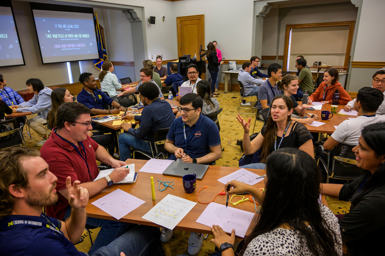 Students sit in groups at tables in a large classroom, chatting with one another, with sheets of paper in front of them.