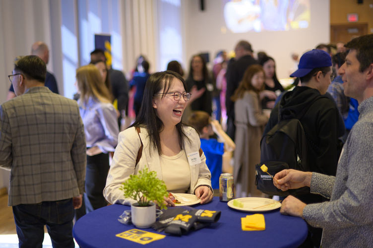 A MADS student laughs at a table while chatting with a faculty member, with a larger reception visible behind them.