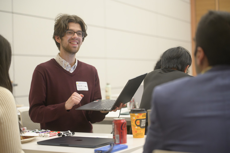 A person holds a laptop while speaking to Neil Tambe at a table. Only the back of Tambe's head is visible