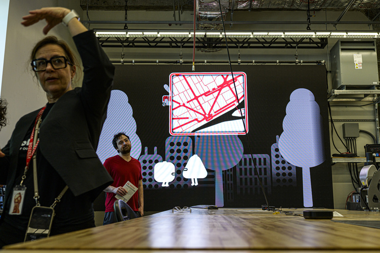 An artist in black clothes gestures with one arm while standing in front of an extended reality exhibit that features a project Detroit stree grid and multiple violet trees