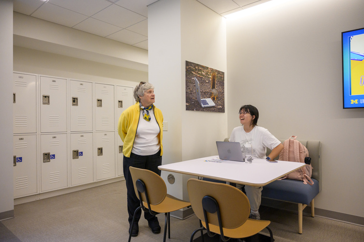 Sue Quackenbush standing beside a table where a student is seated at a laptop, chatting with them.