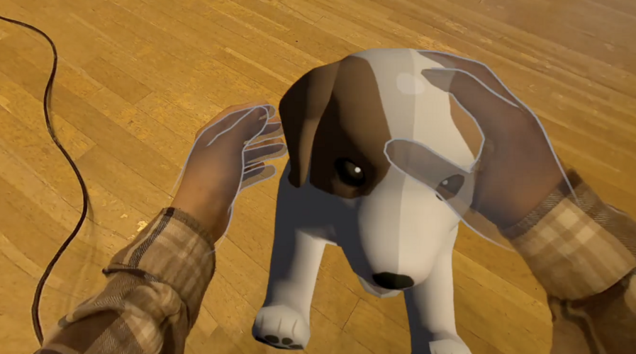 A dog appears on a wooden floor in mixed realty, with hands patting its head