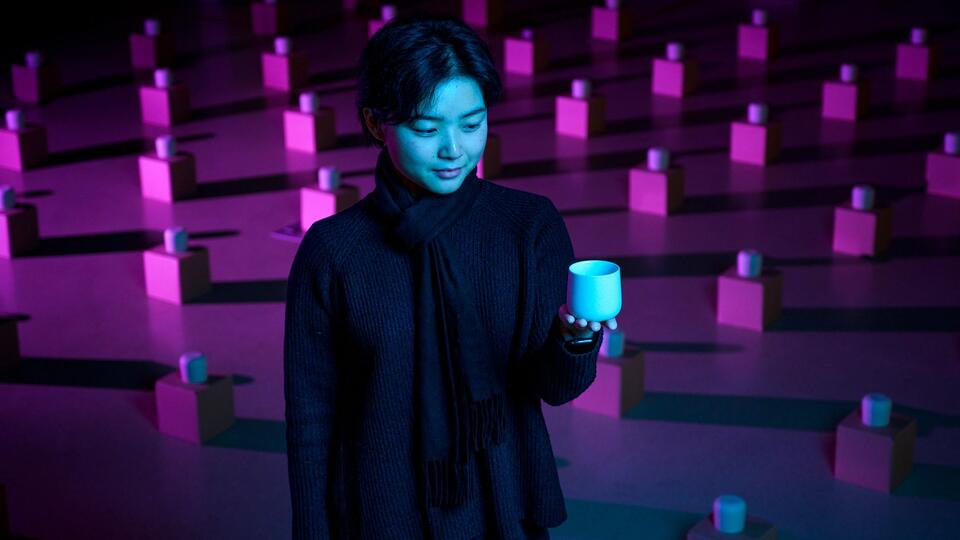 Lynette sits among an art exhibit of many tea cups on boxes laid out in a grid pattern across the floor.