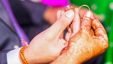 The hands of a bride and groom holding rings. The bride's hand is adorned with intricate henna. 