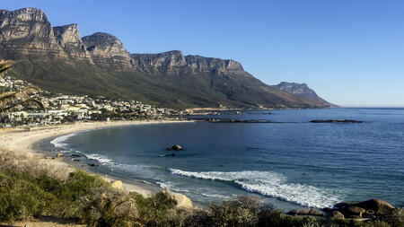 Landscape photo of Cape Town, South Africa coastline. The image captures people on the shore and in the water, and buildings below jagged cliff sides. 
