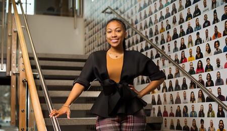 Morgan Miller stands and smiles on a staircase in the Trotter Multicultural Center, in front of a wall decorated with headshots of hundreds of U-M community members.