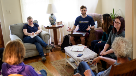 A group of six people sit on armchairs and wooden chairs around a coffee table in a homey environment. One person takes notes in a notebook, and others listen to a person wearing a University of Michigan School of Information T-shirt who gestures openly while balancing a laptop on their lap. 
