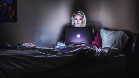 A person sits up in bed and uses a laptop in a dark room.