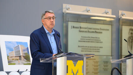 Donor Larry Leinweber speaks at a podium adorned with a Block M. Behind him are commemorative hardhats and a rendering of the Leinweber Computer Science and Information building. He is wearing glasses and a blue blazer. 