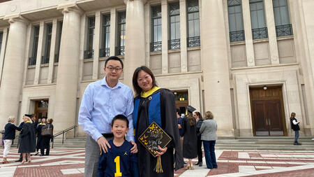 Yao Wei dressed in graduation gown stands outside of an auditorium on University of Michigan campus with her husband and son on her graduation day. She holds a decorated graduation cap.