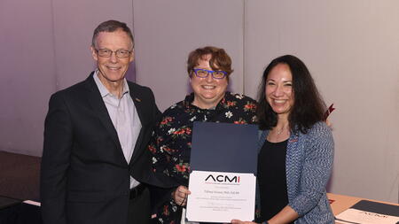 Tiffany Veinot is flanked by the past and present presidents of ACMI. They are smiling and holding an award. 