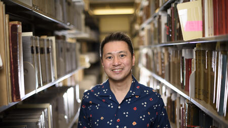 Ricky Punzalan, wearing a blue shirt, smiles as he stands among the stacks of archives in a University of Michigan library. 