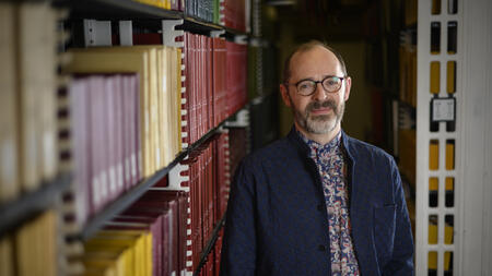 Jesse Johnson wears glasses, a cardigan and stands next to a bookshelf. 