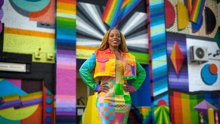 Megan poses in front of a rainbow and prismatic mural in an alleyway while wearing a vibrant dress and jacket of a patchwork of shapes and colors.