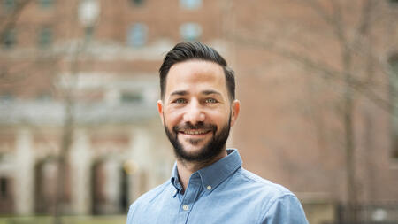 Alain Cohn wearing a button up blue shirt, standing outside, and smiling at the camera. 