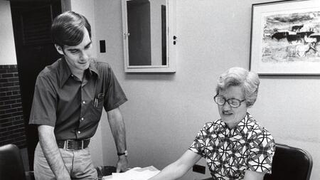 An archival photo of former UMSI professor Constance Rinehart. She is shown sitting at a desk and holding a document. A young man is looking over her shoulder. 