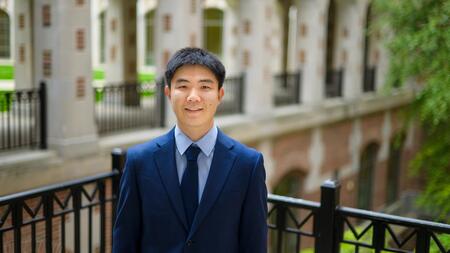 Jiaxin Pei wearing a blue suit and tie, standing outside and smiling at the camera. 