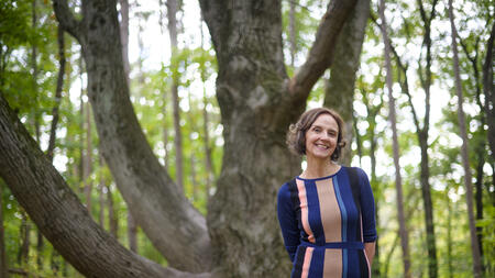 Nicole Ellison in front of a tree on a hiking trail, wearing a blue and pink dress and smiling.  