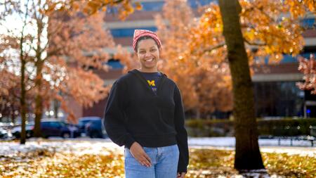 An image of Christine Carethers standing on campus surrounded by fall foliage, wearing a U-M t-shirt and a red bandana