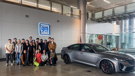 An image of students posing beside a classic Cadillac model in the lobby of General Motors' Cole Building, with an illuminated "gm" sign on the wall behind them